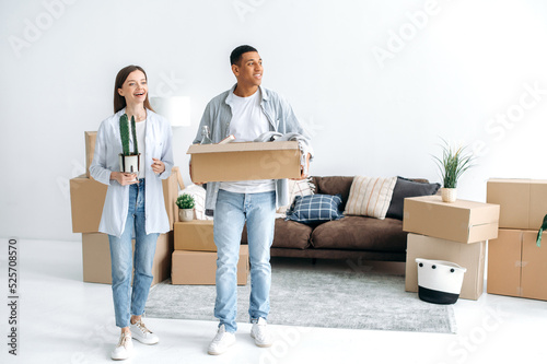 Moving to a new apartment. Happy multiracial young couple in love walk into their new home, holding boxes of household items, look around the interior with delight, rejoice at their new home, smile