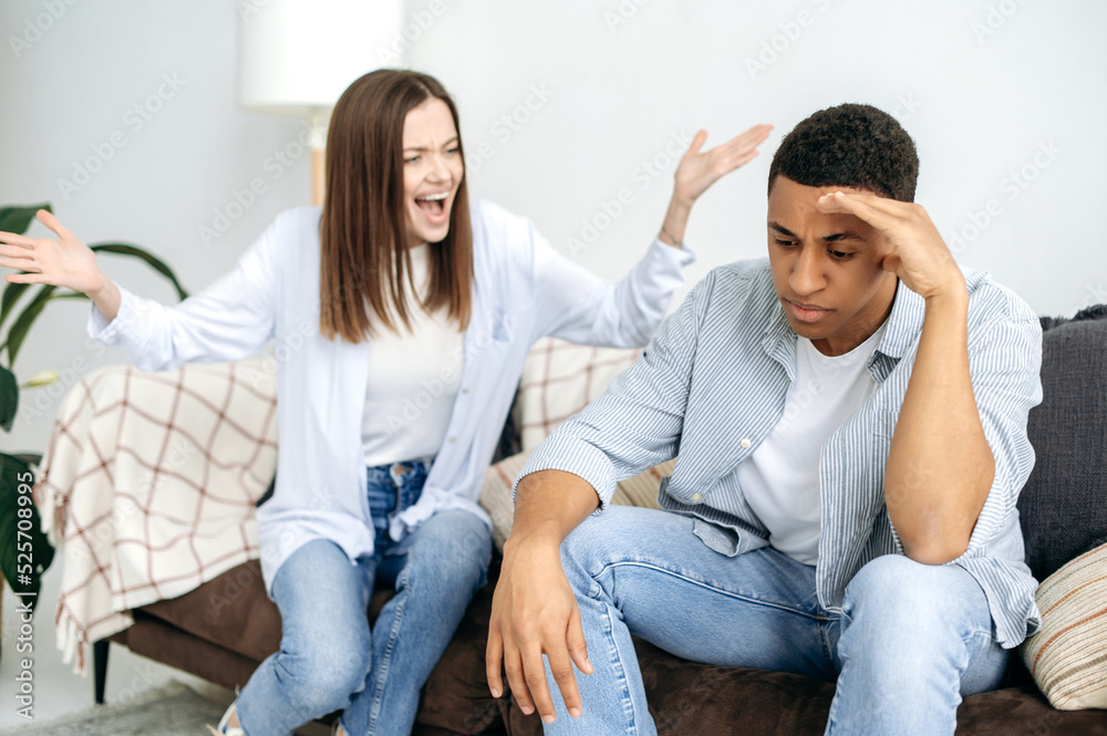 Misunderstanding in relationships, jealousy, quarrel. A married multiracial couple sorts things out while sitting in the living room on a sofa, a caucasian woman screams at her hispanic husband Misunderstanding in relationships, jealousy, quarrel. A married multiracial couple sorts things out while sitting in the living room on a sofa, a caucasian woman screams at her hispanic husband