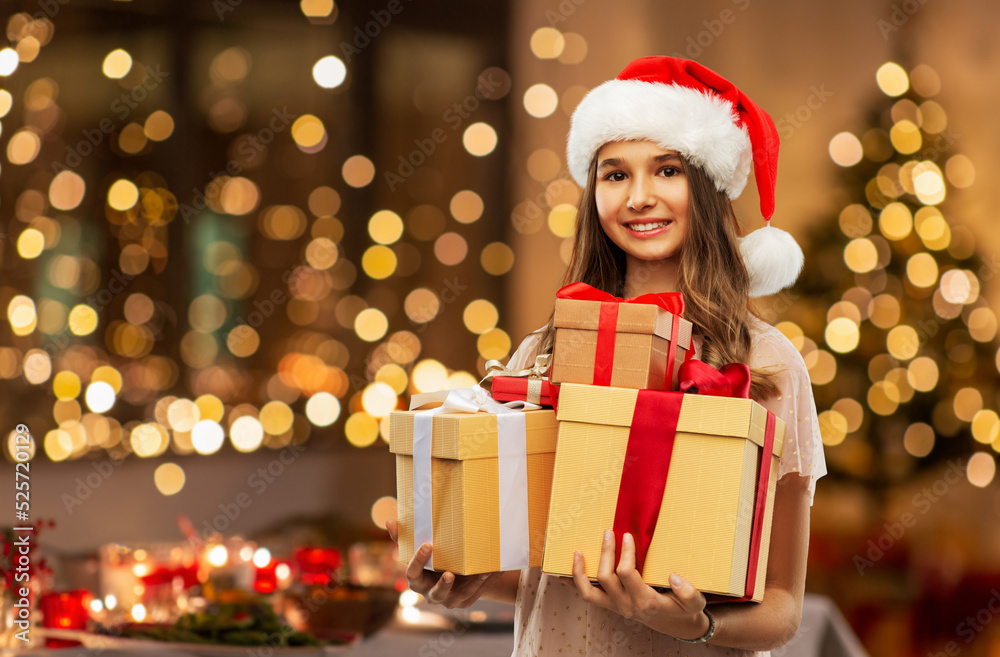 christmas, holidays and people concept - happy smiling teenage girl in santa helper hat holding gift box over festive lights at home on background christmas, holidays and people concept - happy smiling teenage girl in santa helper hat holding gift box over festive lights at home on background
