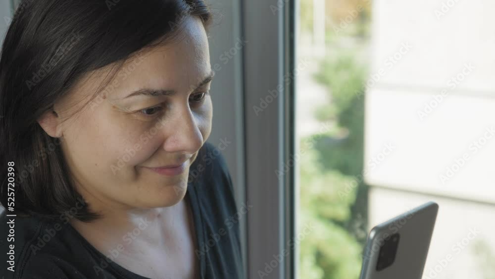A girl stands near the window in a room with a phone in her hands, smiles and looks at the phone. Outside the window you can see the city, with houses, trees and a road along which cars drive. A girl stands near the window in a room with a phone in her hands, smiles and looks at the phone. Outside the window you can see the city, with houses, trees and a road along which cars drive.