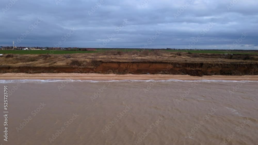 Motion along deserted sand beach under grey sky with heavy clouds. Water washes sand with foamy waves against dry grass meadow in winter aerial view from sea Motion along deserted sand beach under grey sky with heavy clouds. Water washes sand with foamy waves against dry grass meadow in winter aerial view from sea