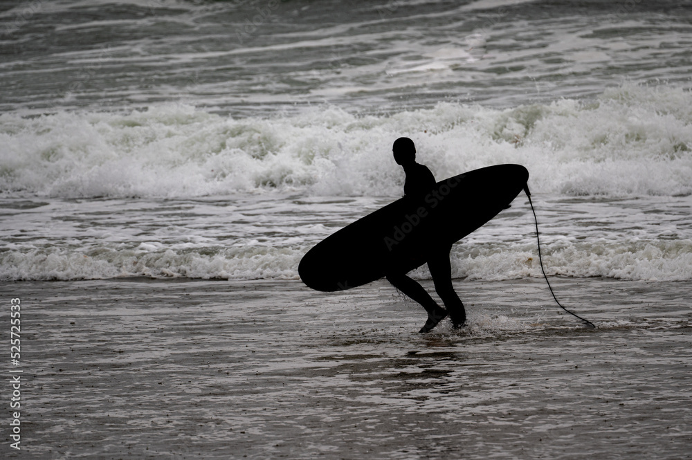 Silhouette of a male surfer walking along the beach with his surfboard with waves in the background