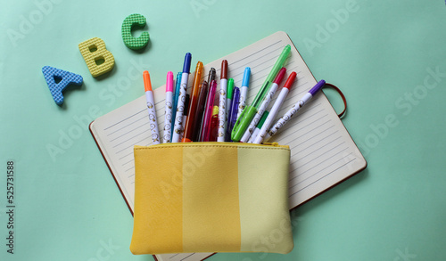 Bolsa amarilla con plumones de colores sobre cuaderno de rayas y letras de goma del abecedario en colores azul, amarillo y verde. 