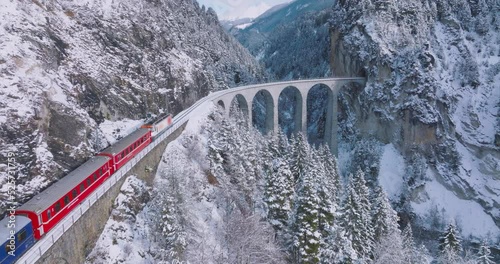 Landwasser Viaduct world heritage sight with luxury Glacier and Bernina express in Swiss Alps snow winter scenery. Aerial Drone shot red train passing through famous mountain in Filisur, Switzerland. photo