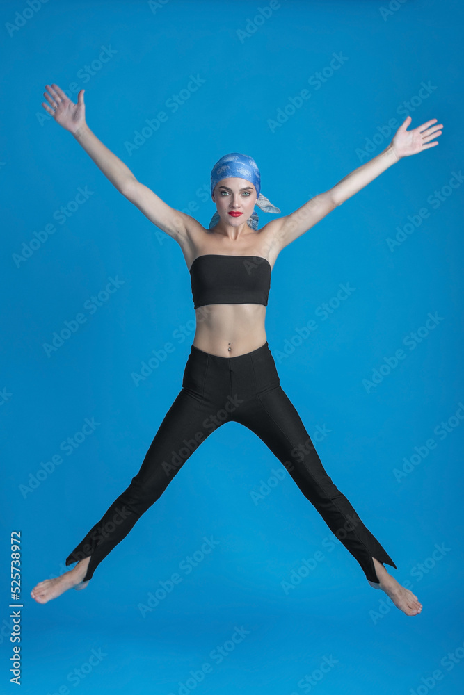 Obraz premium Sport, fashion and make-up concept. Slim and beautiful woman with black and tight sport outfit, scarf and red lipstick jumping in air. Graceful pose freeze in motion. Blue studio background