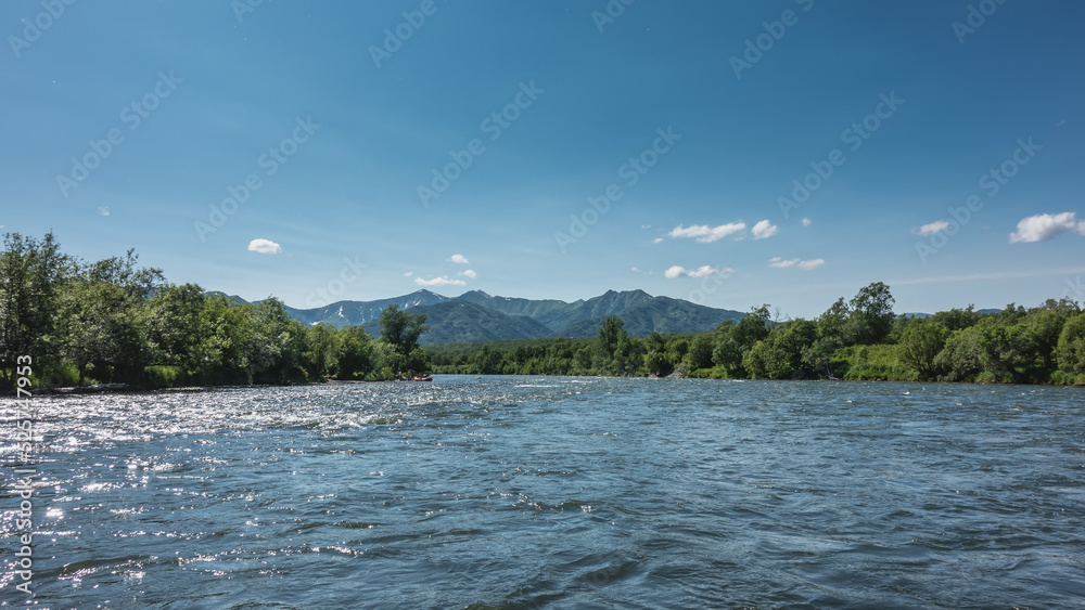 The blue river sparkles in the sun. There is lush green vegetation on the banks. A mountain range against the sky. Kamchatka. River Bystraya