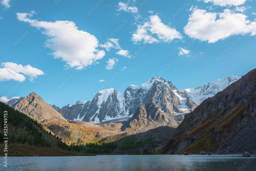 Pure azure alpine lake with view to forest hill and giant snow mountains in autumn sunny day. Glacial lake against huge snow covered mountain range in bright sun. Vivid autumn colors in high mountains