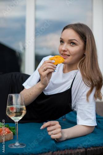 Portrait of young woman lying on blue blanket near glass of white wine, fruits on wooden tray, eating slice of pepper.