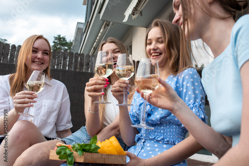 Portrait of young funny beauteous women holding cut vegetables, clinking glasses of white wine, sitting near house.