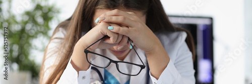 Tired woman doctor sitting at table and holding glasses in clinic photo