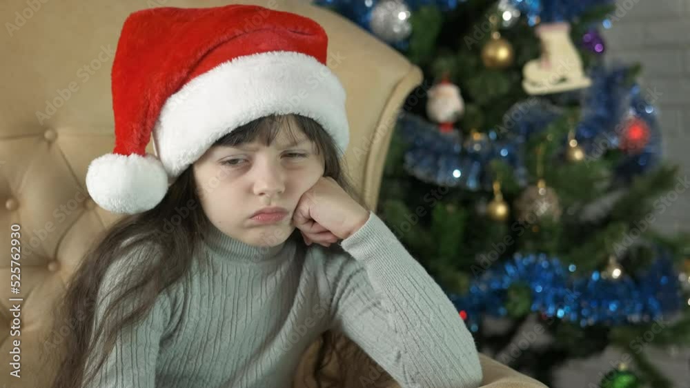 Disappointed during Christmas. An upset child in Santa hat with disappointed sits in the armchair by the Christmas tree. Disappointed during Christmas. An upset child in Santa hat with disappointed sits in the armchair by the Christmas tree.