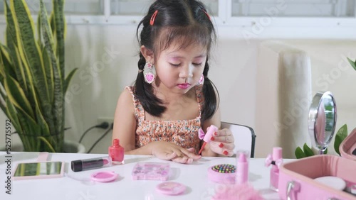 Asian adorable funny little girl making makeup dips brush into bottle to paints nails polish red nail varnish herself, Learning activity to be woman, happy kid is beautiful make up with cosmetics toy photo