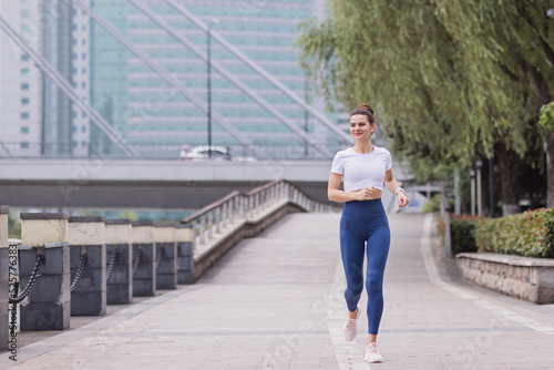 Young brunette Woman running on asphalt road in summer park. Active sporty caucasian female has morning workout. Athletic person in sportswear exercises outdoor. Dressed leggings and sportive top