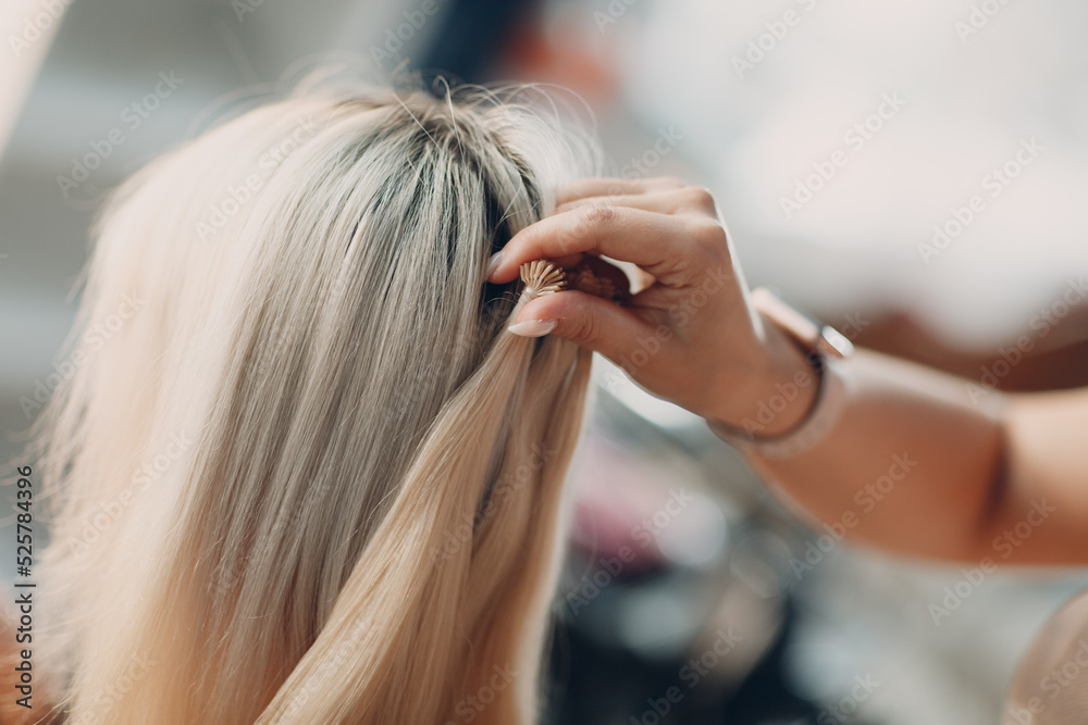 Fototapeta premium Hairdresser female making hair extensions to young woman with blonde hair in beauty salon. Professional hair extension