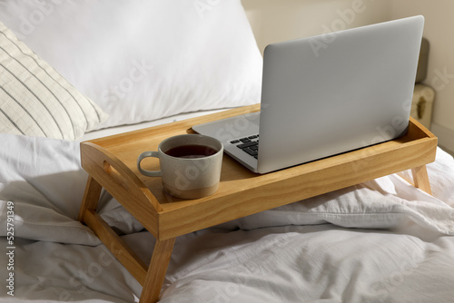Wooden tray with modern laptop and cup of aromatic tea on bed indoors