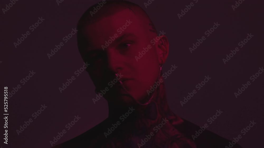 Chest up shot of tattooed man with ear to nose chain looking at camera and moving his head to music in dark studio with blinking neon light projection Chest up shot of tattooed man with ear to nose chain looking at camera and moving his head to music in dark studio with blinking neon light projection