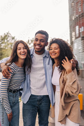 Three young interracial students with good mood are standing in embrace outdoors. Brunettes guy and girls wear casual clothes. Happy day concept