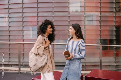 Two young interracial girls are friendly talking to each other standing outdoors. Brunette female students wear casual clothes. Lifestyle concept