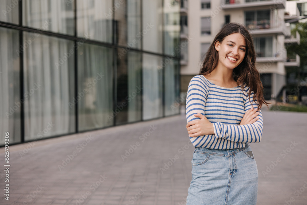 Smiling young caucasian girl with arms crossed in front of her standing outdoors. Brunette woman with wavy hair wears casual clothes. Relaxation concept