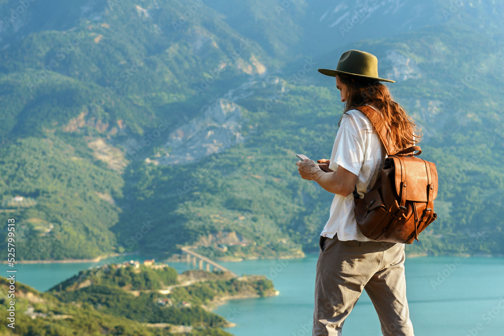 Traveler man with hat and backpack holding smartphone against scenic view of lake and mountains