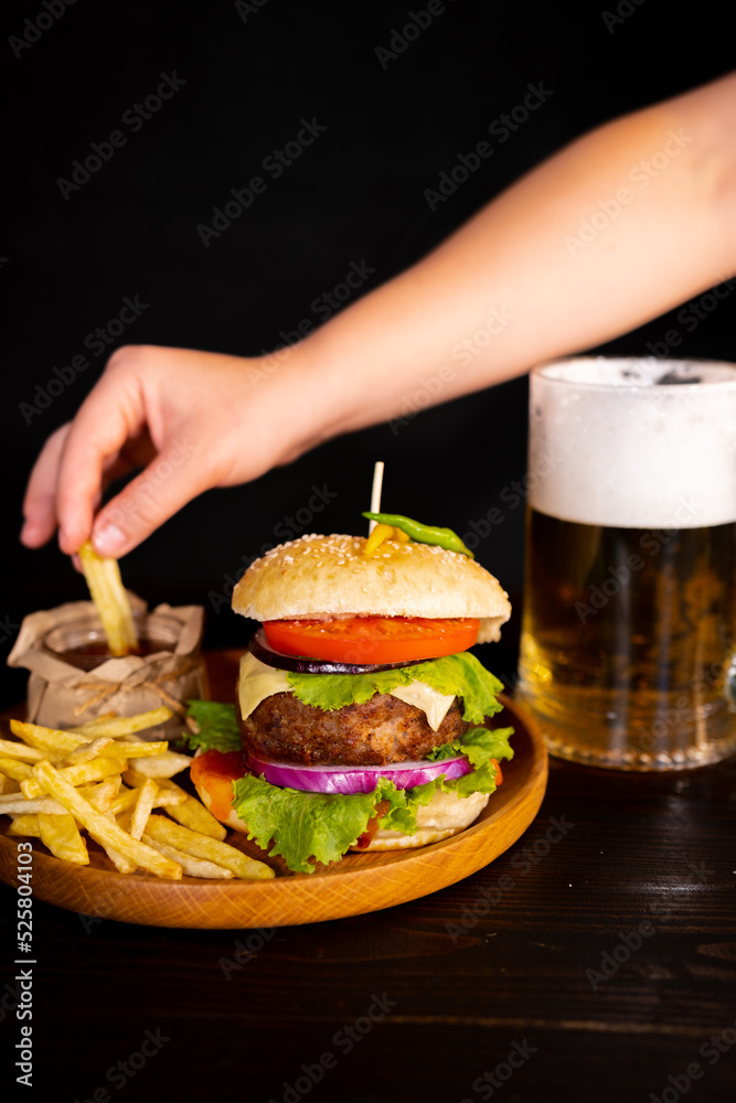 tasty hamburger burger sandwich with french fries and ketchup on a wooden tray a glass of cool beer with foam fast food black background. hand dips french fries in ketchup