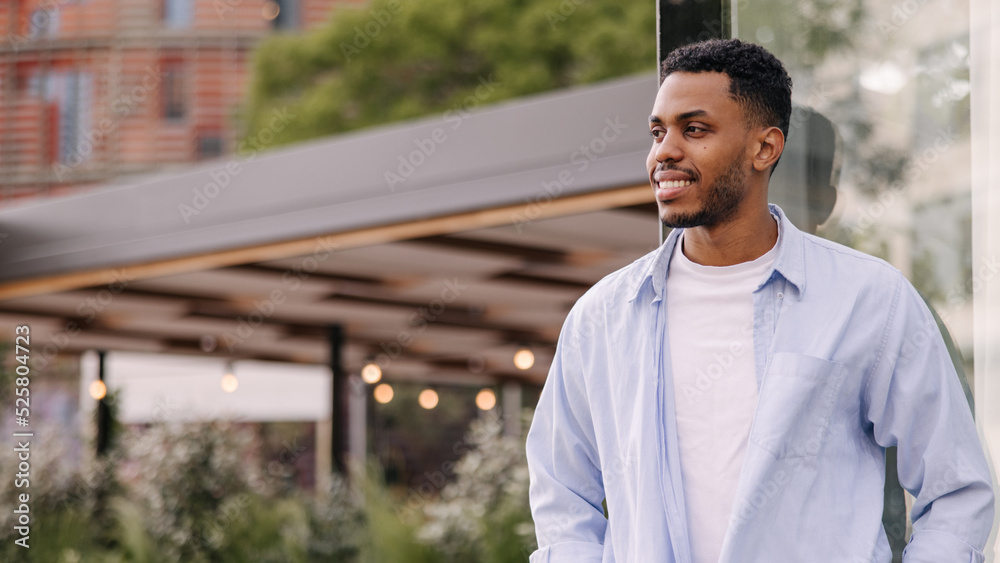 Fototapeta premium Handsome young african guy is looking towards city relaxing outdoors. Brunette man with stubble wears t-shirt and shirt. Lifestyle concept