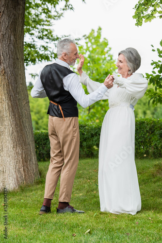 happy mature man in formal wear dancing with bride in white wedding dress in green garden
