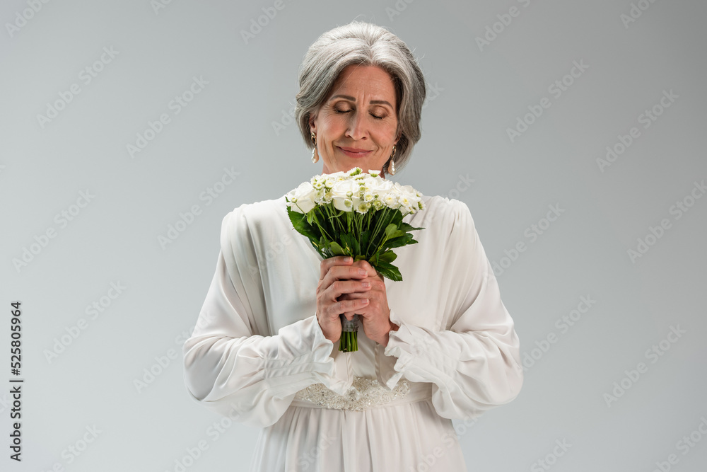 joyful middle aged bride in white wedding dress holding bouquet and smelling flowers isolated on grey joyful middle aged bride in white wedding dress holding bouquet and smelling flowers isolated on grey