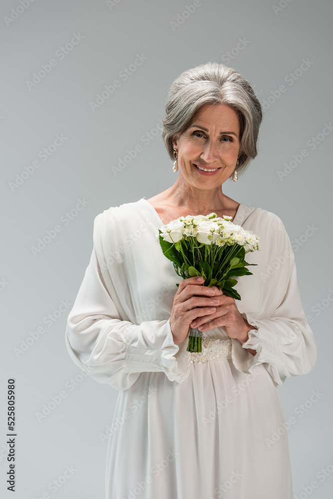 cheerful middle aged bride in white wedding dress holding bouquet isolated on grey cheerful middle aged bride in white wedding dress holding bouquet isolated on grey