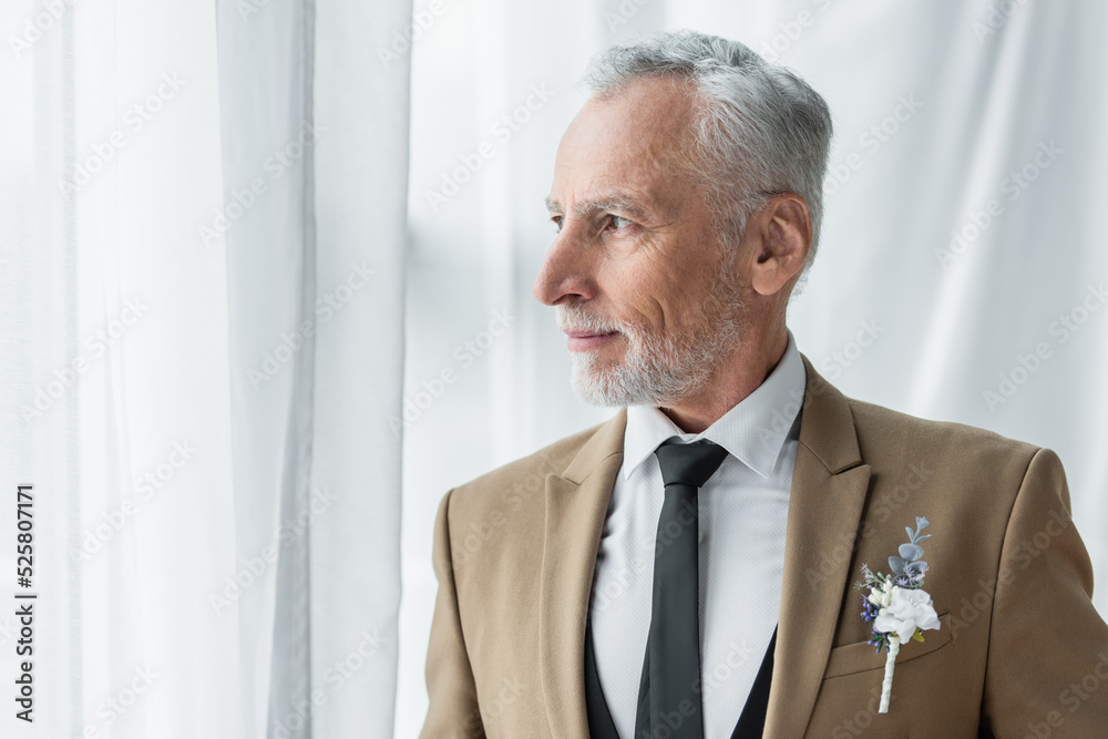 bearded middle aged man in suit with boutonniere looking away near white curtains bearded middle aged man in suit with boutonniere looking away near white curtains