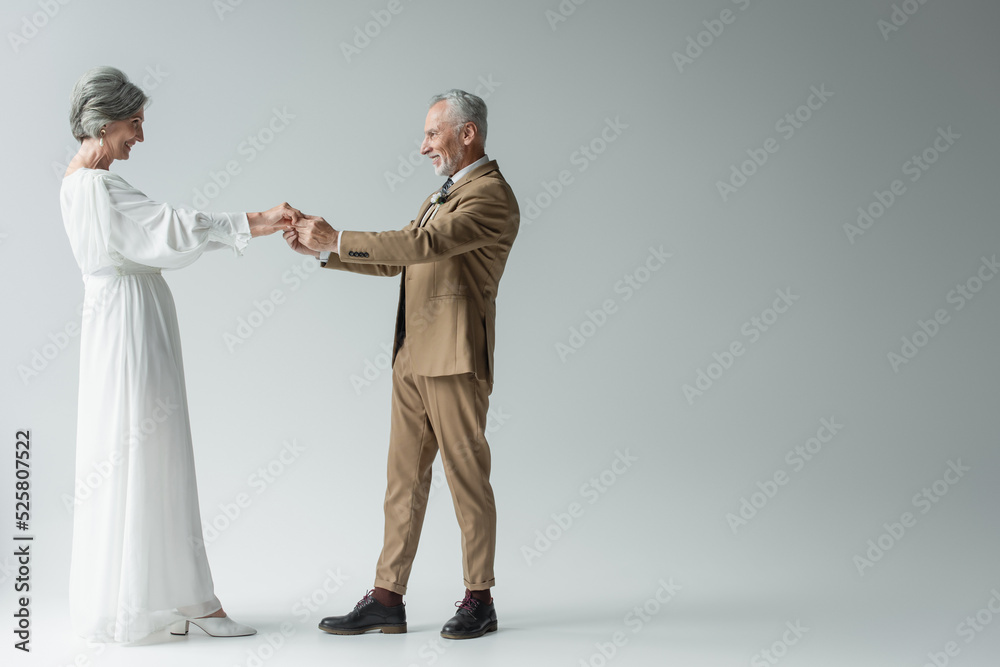full length of cheerful middle aged man in suit and bride in white wedding dress holding hands on grey full length of cheerful middle aged man in suit and bride in white wedding dress holding hands on grey
