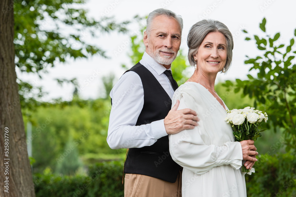 mature man in formal wear hugging smiling bride with wedding bouquet in garden mature man in formal wear hugging smiling bride with wedding bouquet in garden