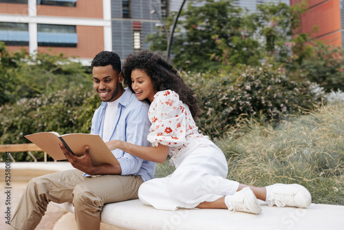 Nice african young friends reading essay preparing for lessons sitting outdoors. Brunette guy and girl spend their leisure time studying, wear casual clothes.