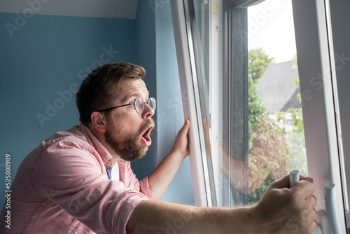 Shocked, frightened man looks out the window and screams excitedly, standing against the background of the blue wall in the room. 