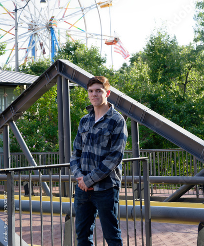 Handsome young man in front of iron railing and looking confident with a ferris wheel in the background.