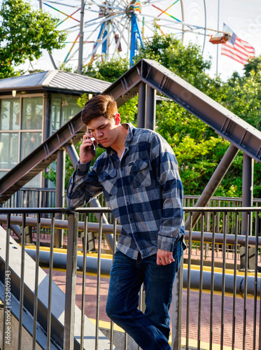 Handsome young man standing in front of iron railing and talking on a cellphone with a ferris wheel in the background.