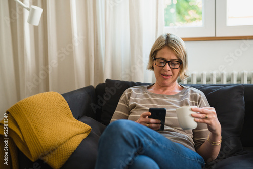 caucasian woman sits on sofa in her living room drinking coffee and texting on mobile phone © cherryandbees