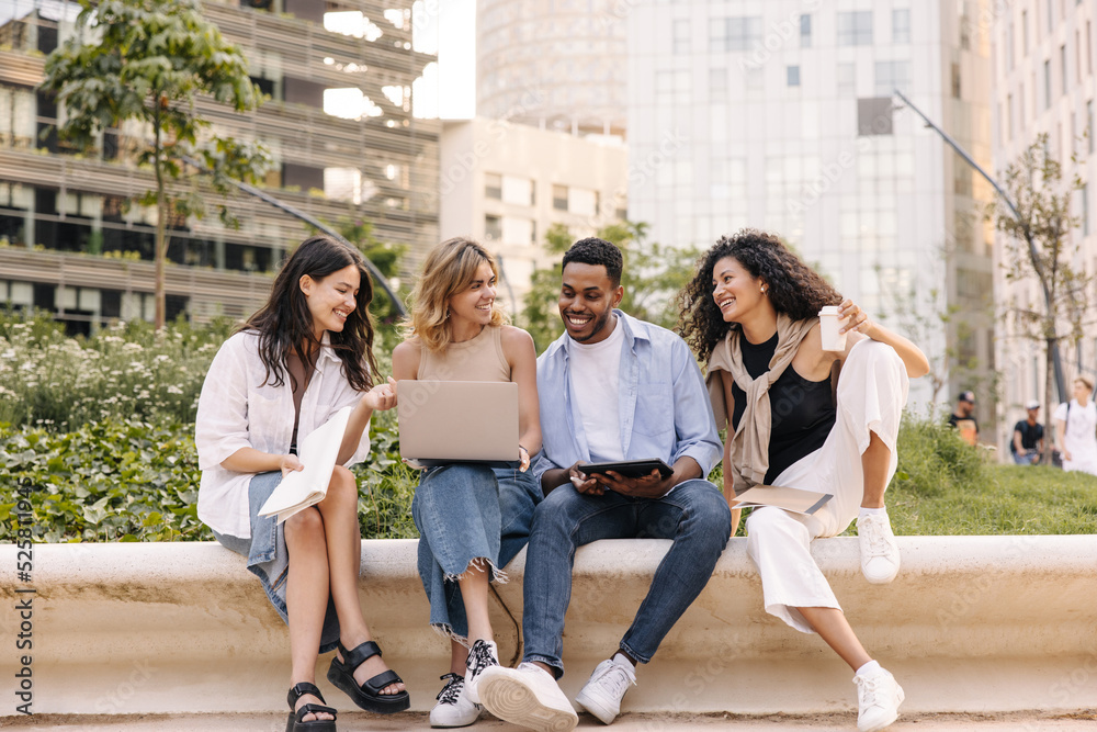Fototapeta premium Four successful young interracial students discuss new topic among themselves relaxing sitting on campus. Guy and girls with laptop, tablet and notebook. Concept of learning