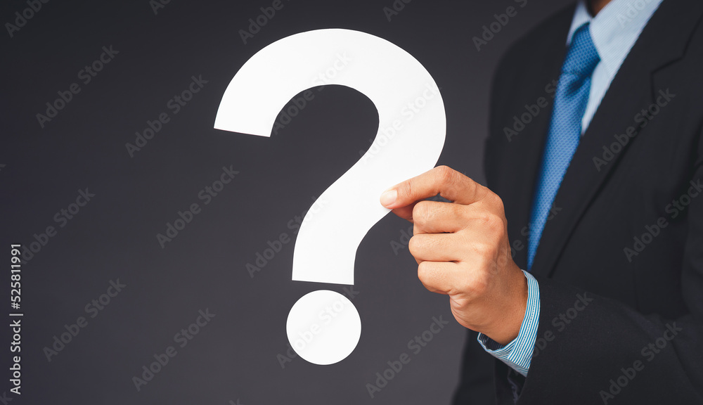 Businessman in a suit holding a white question mark paper while standing on a gray background Businessman in a suit holding a white question mark paper while standing on a gray background
