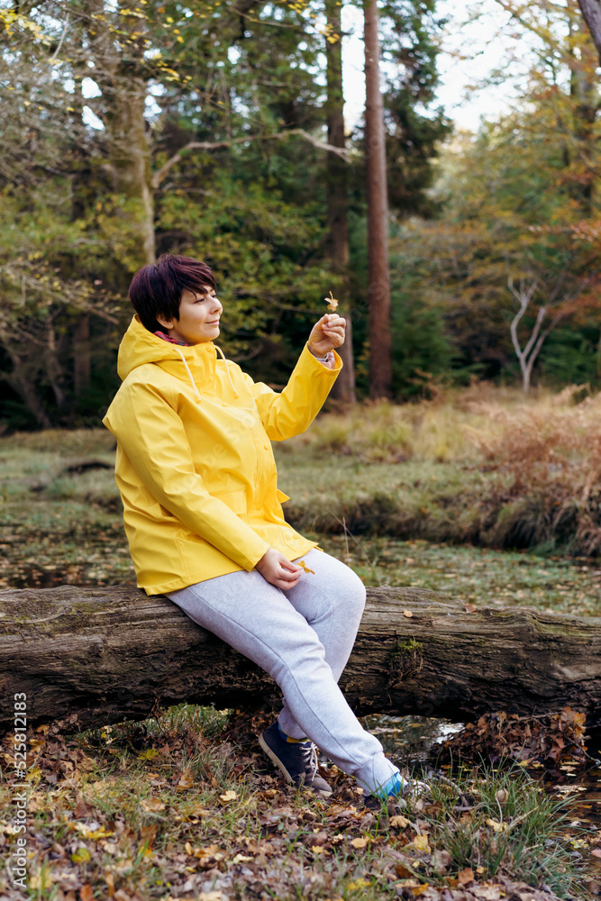 Woman in bright yellow raincoat sitting on a fallen tree with fallen leaves and enjoying the moment in the forest. Feeling harmony, reunion with nature in autumn. Relaxing, personal fulfillment.