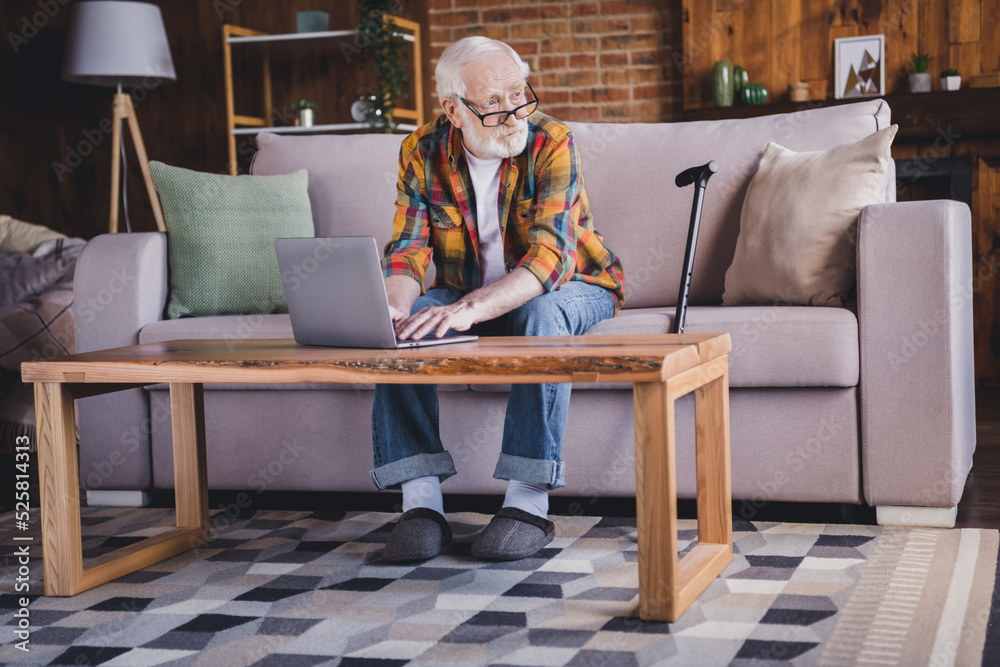 Photo of busy thoughtful man pensioner wear checkered shirt chatting modern gadget sitting sofa indoors house room