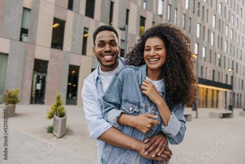 Happy young african guy hugging his girlfriend from behind looking at camera standing on street. Brunette with curly hair wears denim jacket. Good mood concept