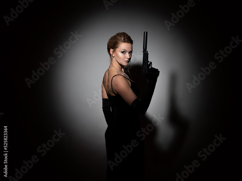 A dangerous spy. A femme fatale in a black dress with an open back holds a pistol with a silencer in her hands, a portrait in a spot of light