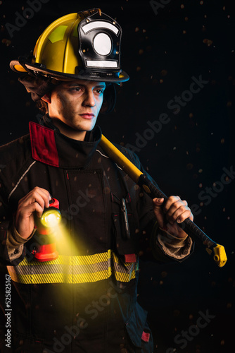 Fireman standing, holding hammer, wearing special protective uniform, with lighted flashlight.  Black background.  Night and fire.