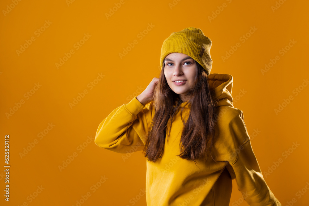 a young cheerful brunette in bright clothes is dancing and having fun. A young woman in a yellow hoodie and a hat on a yellow background a young cheerful brunette in bright clothes is dancing and having fun. A young woman in a yellow hoodie and a hat on a yellow background