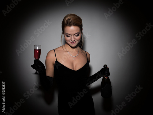 beautiful woman in a black dress with a neckline and long velvet gloves, dancing and holding champagne in her hands. Attractive young blonde with makeup for a celebration, portrait in a spot of light