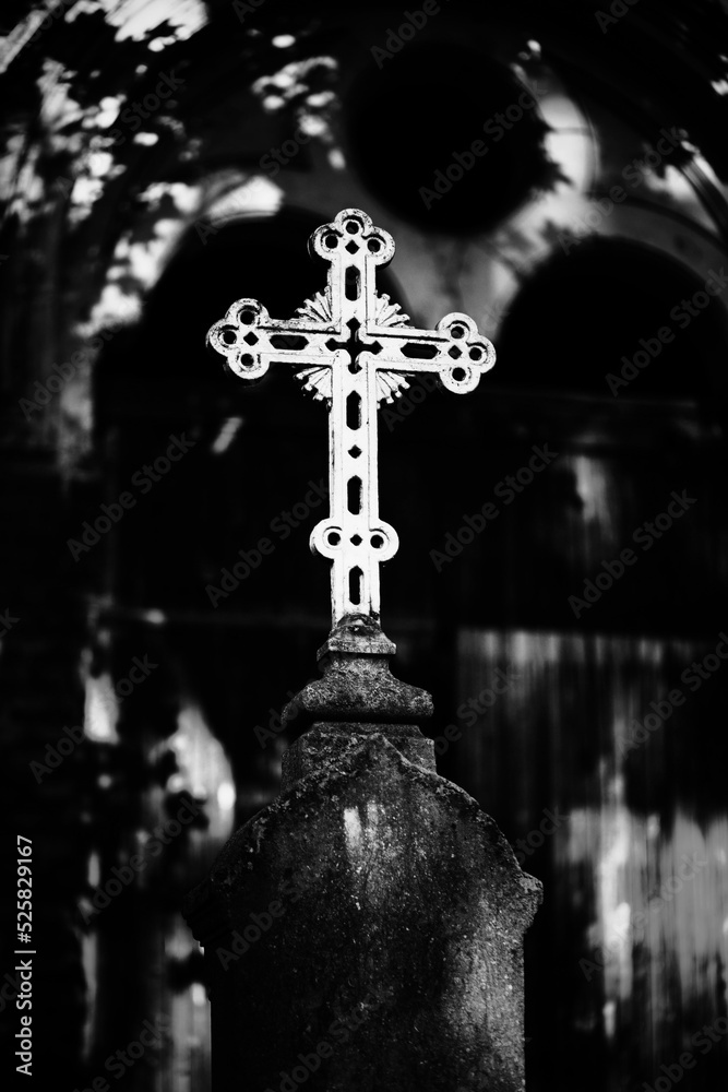 A black and white image of a white Catholic old cross standing on a pedestal in the cemetery near the crypt. Religion and death.