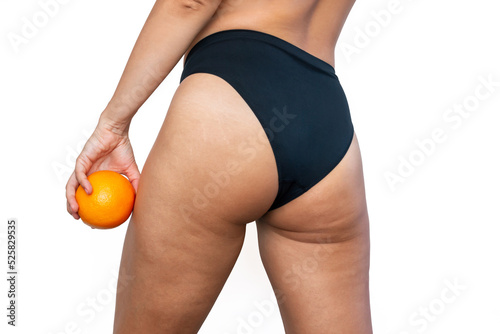 Cropped shot of young woman with cellulite and white stretch marks from a weight loss or weight gain on thighs holding an orange in her hand isolated on a white background. Excess weight, overweight