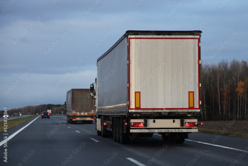 Semi trucks convoy drive on countryside asphalted road at autumn evening. Goods delivery logistics in Europe