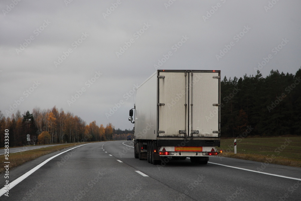 Heavy white semi truck back view on two-lane countryside asphalted road at autumn evening on gray cloudy sky background